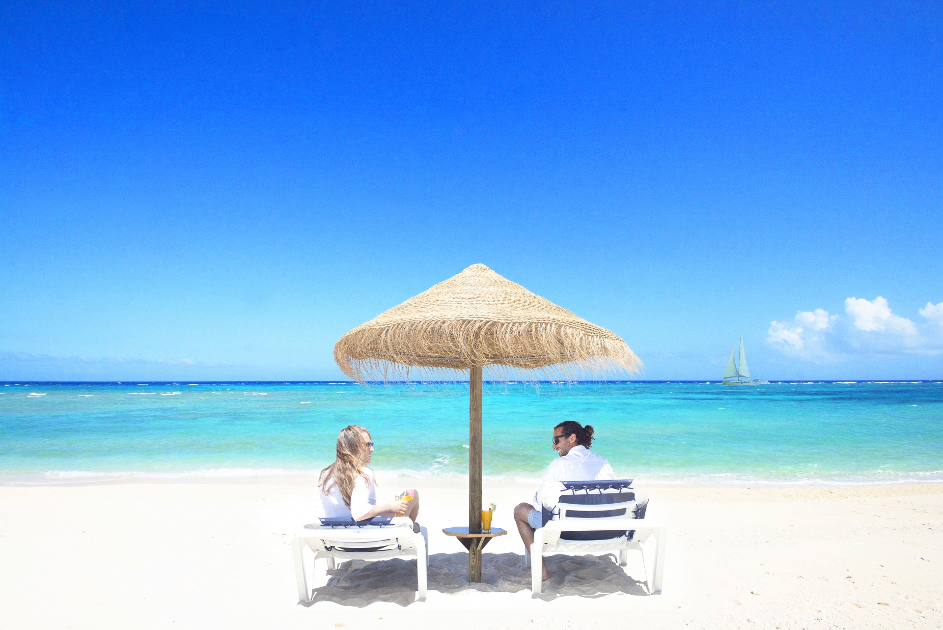 A couple sitting on beach chairs on the sand at the beach, under a straw sunshade