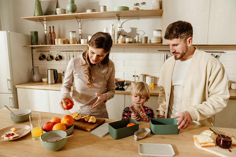 Mother stands at the table holding an apple while her son stands next to her holding the lid of a lunch box held by his father, in the kitchen.