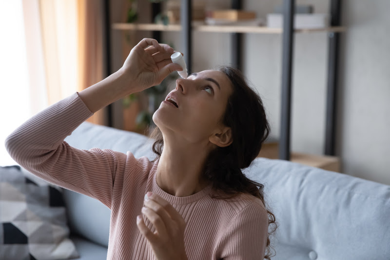 Woman in pink top tilting her head back, and using eye drops on her right eye.