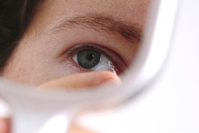 Woman inserting contact lens into her right eye, seen through the reflection of a handheld mirror