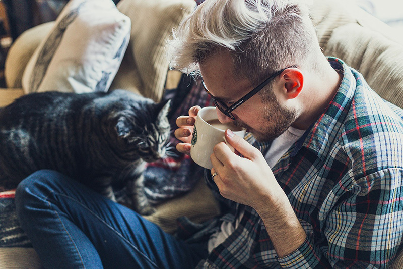 An image of a man with glasses drinking coffee with his cat