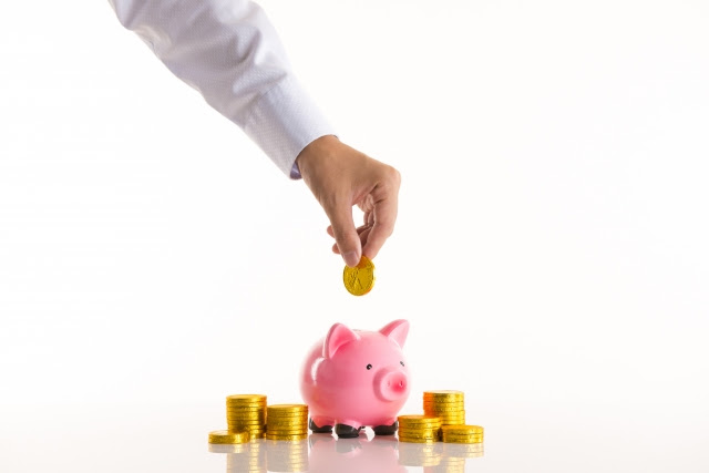 Arm in white sleeve dropping gold coin into pink piggy bank, surrounded by more gold coins