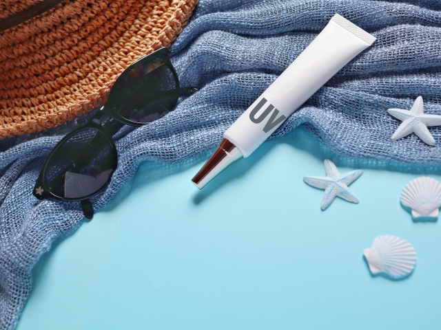 Blue towel, black sunglasses, straw hat, white unbranded sunscreen and seashells.