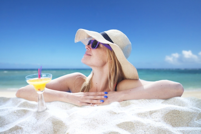 Woman wearing a hat and sunglasses, at the beach, with a yellow drink in a cocktail glass