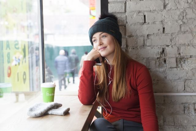 Redhead woman sitting at a cafe with a takeaway cup and gloves, looking out the window, showing the city and people