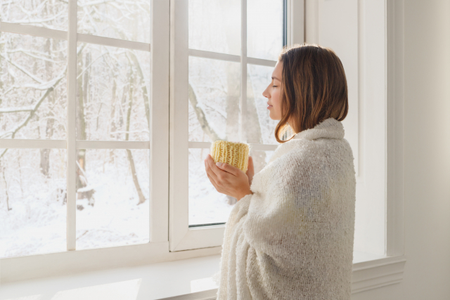 Brunette woman wrapped in white blanket holding tea cozy, and looking out the window, looking at a winter forest