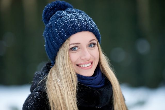 Medium close up of blonde woman wearing navy beanie and dark jacket and scarf, looking at the camera, smiling